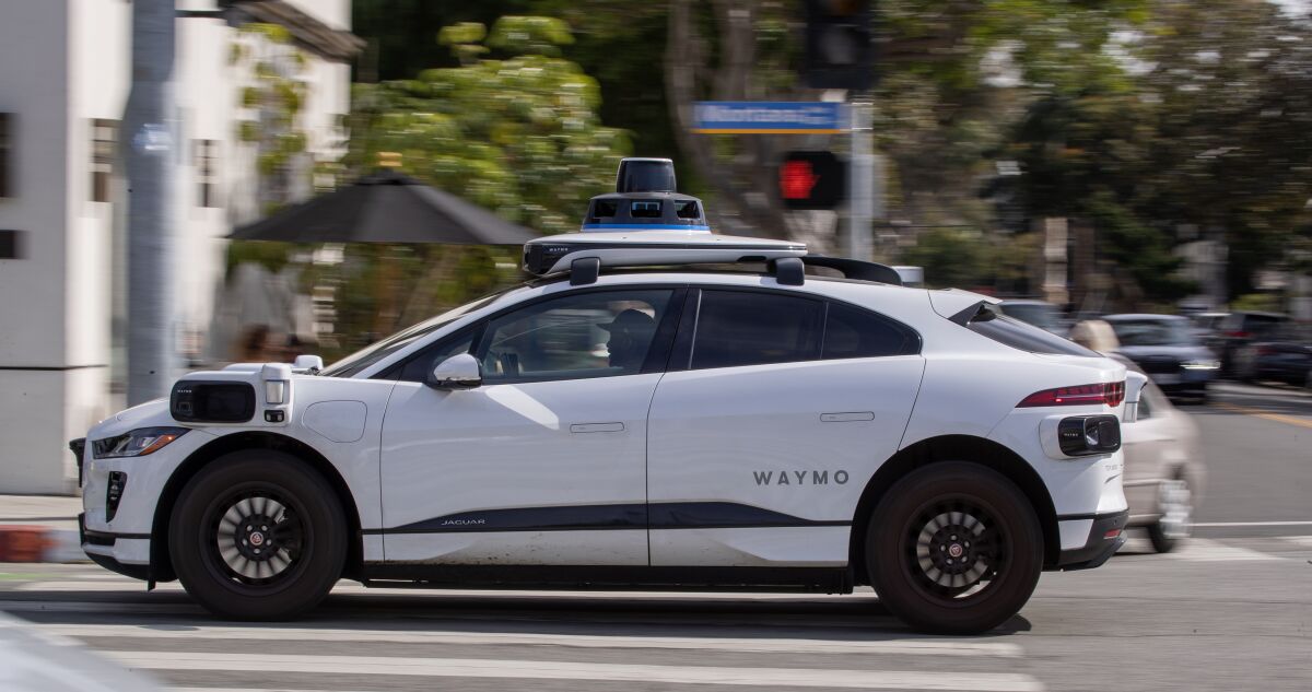 A white car with the word "Waymo" on its side and a self-driving sensor on its roof goes through an intersection.