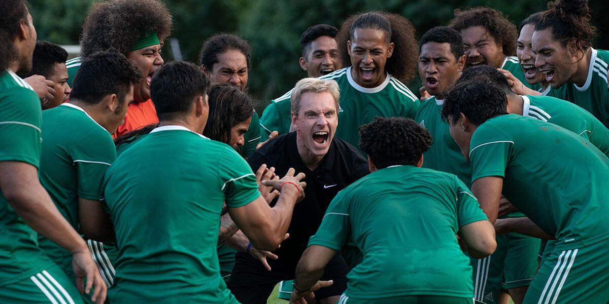 A soccer coach screaming while encircled by players in green jerseys.