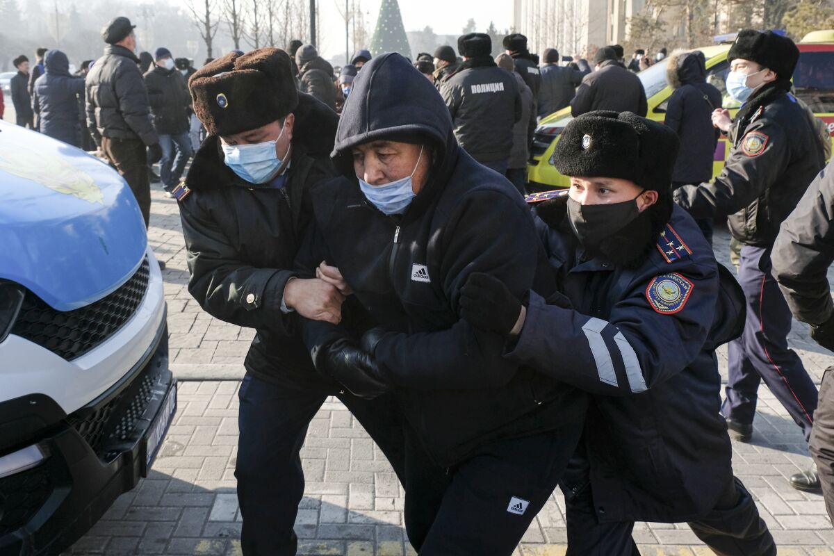 Two police officers in masks and fur hats walk quickly, holding a third man between them by the arms.