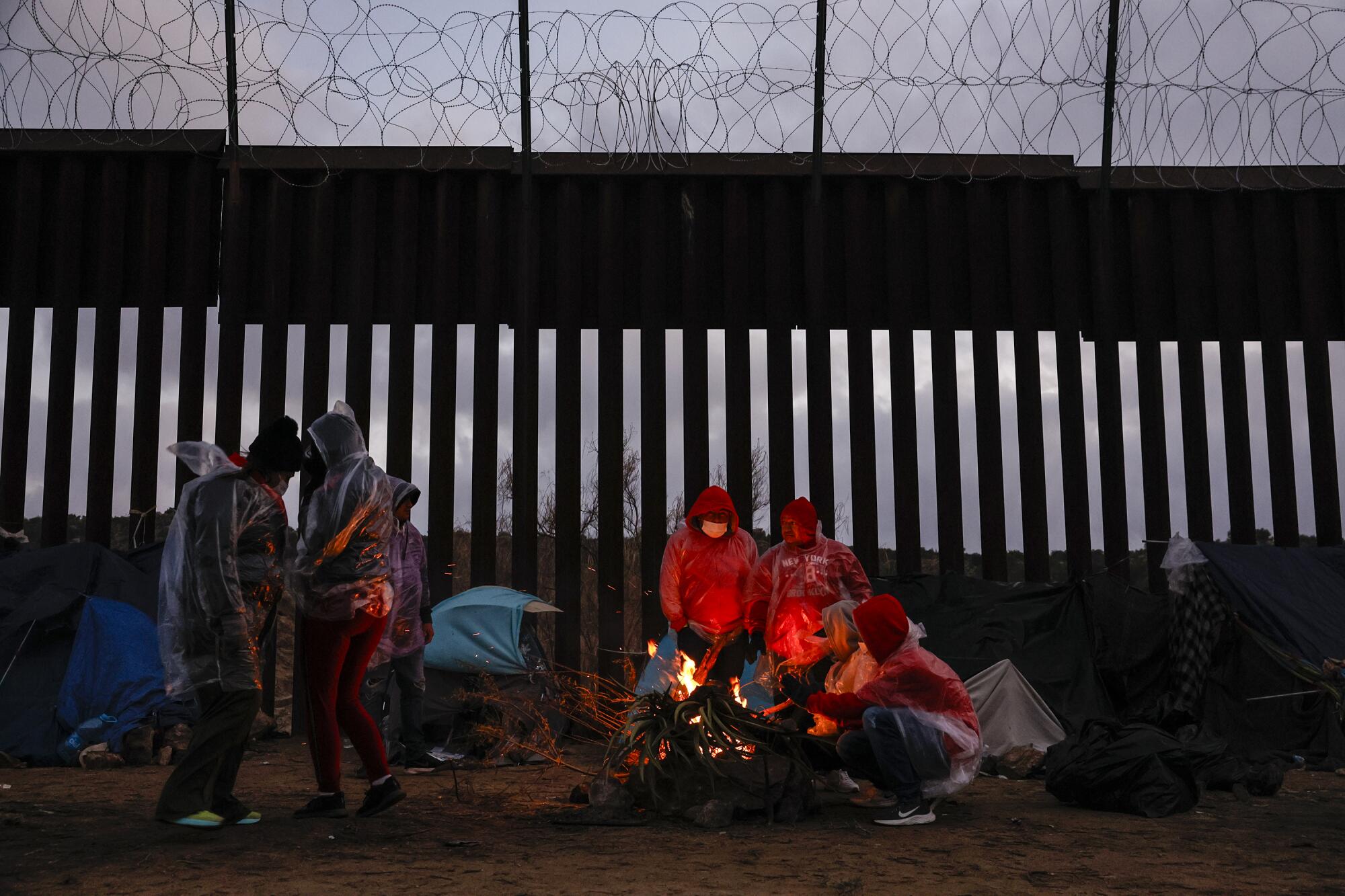 Several people, many in plastic raincoats, next to a barbed-wire-topped wall, a few pacing as others huddle around a campfire