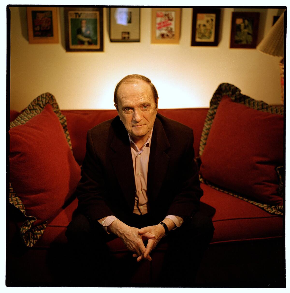 Bob Newhart sitting on a red sofa with his hands together wearing a black suit and open-collar shirt