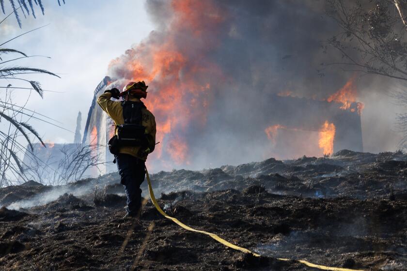 SAN BERNARDINO, CA - AUGUST 5, 2024:A firefighter shields his face from the heat of a fully engulfed home while battling the Edgehill fire on August 5, 2024 in San Bernardino, California.(Gina Ferazzi / Los Angeles Times)