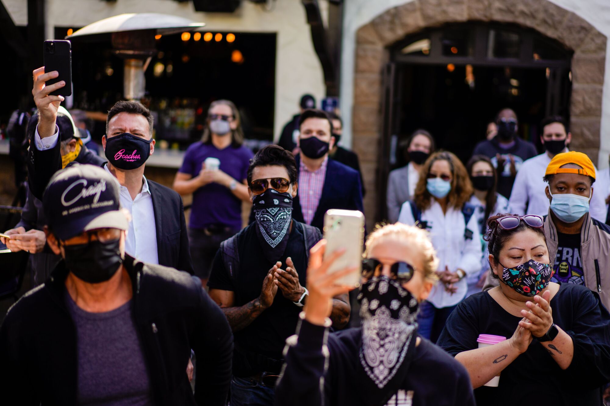 People in bandannas and masks hold up phones and clap at an outdoor news conference.