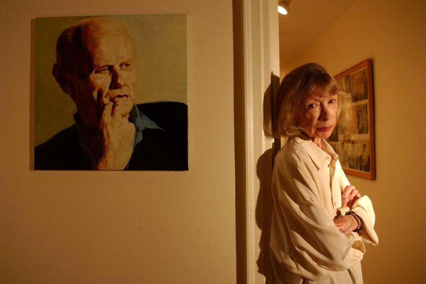 Joan Didion, wearing a white shirt, stands in the hallway of her apartment, next to a portrait of her late husband