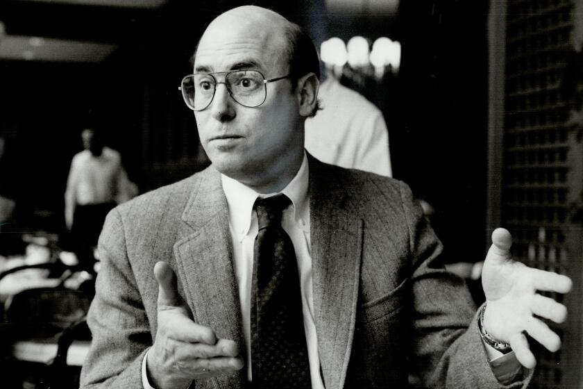 A black-and-white photo of a bald man with glasses in a suit and tie. He has his hands in an expressive position