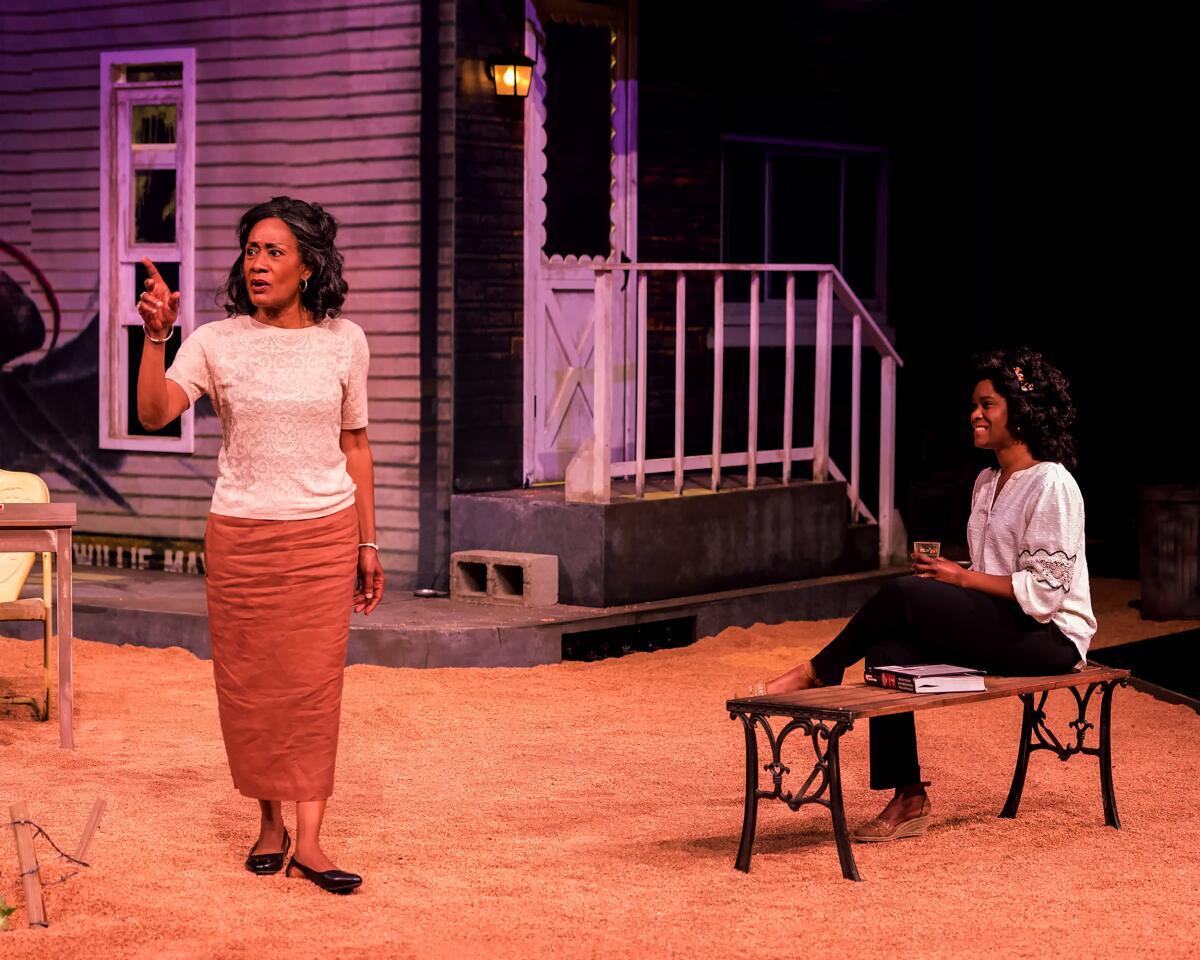 A woman seated on a bench watches a standing woman talking on a stage set representing a backyard outside a house