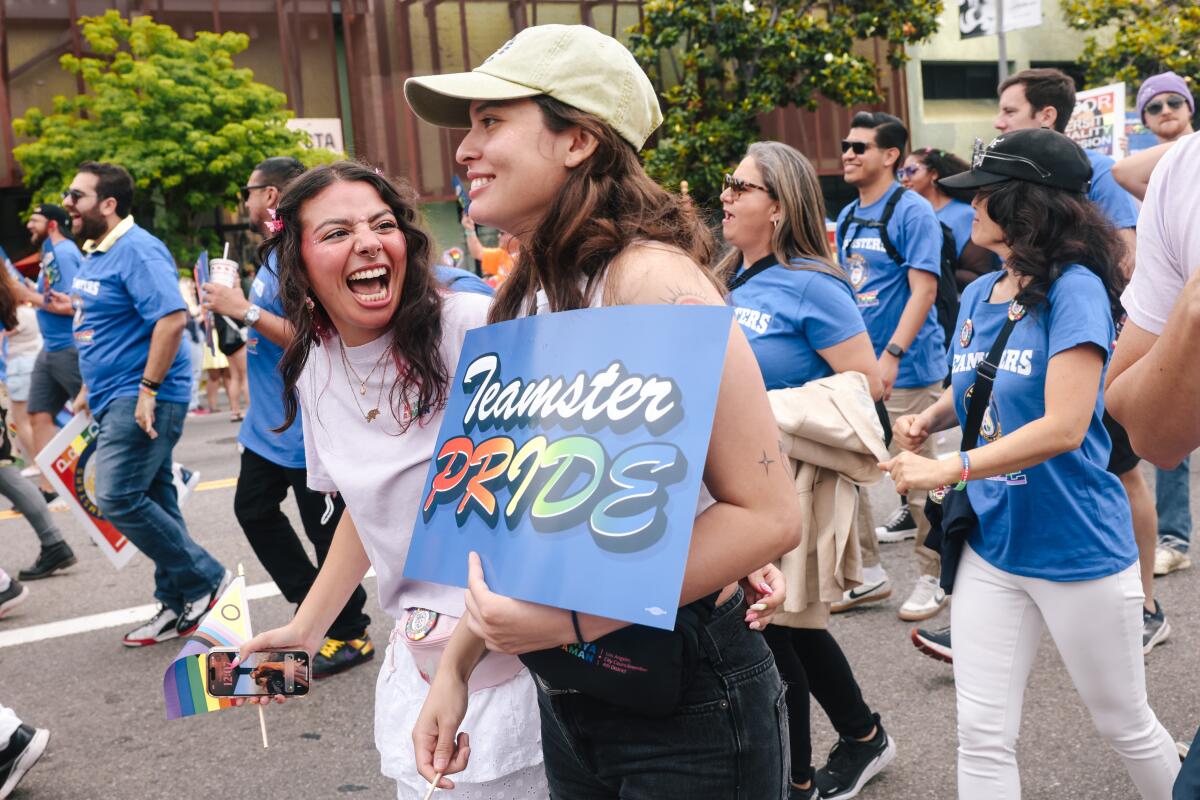 A woman wearing a yellow baseball cap smiles while marching in a parade and holding a rainbow "Teamsters Pride" sign.