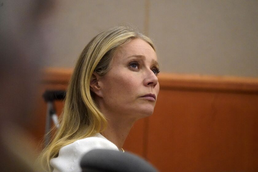Side angle of woman, Gwyneth Paltrow with long blonde hair and a white top, sitting in courtroom looking right