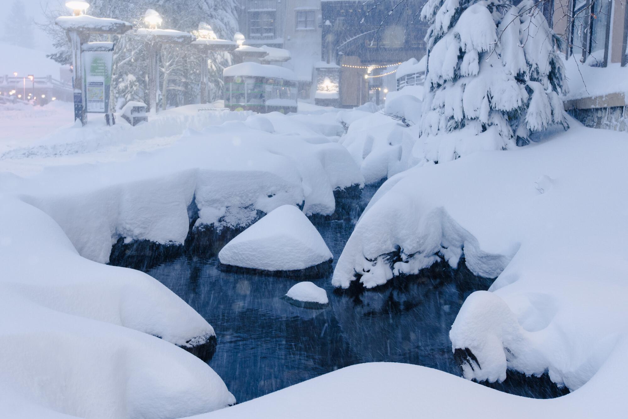 A thick layer of snow surrounds a small body of water near the houses in the background.