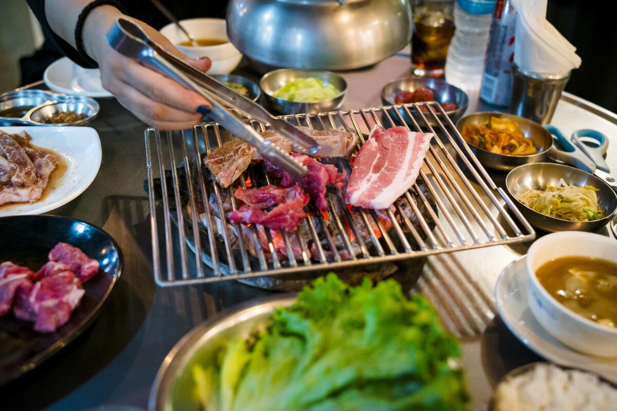 A hand reaches out to cook meat on a tabletop grill, as other ingredients crowd the table's surface.