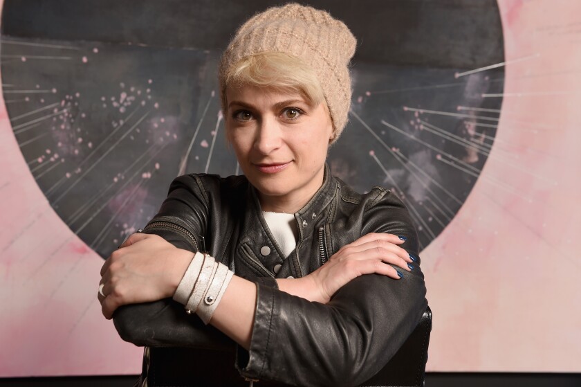A woman in a black jacket and tan knit cap folds her arms in front of a backdrop.