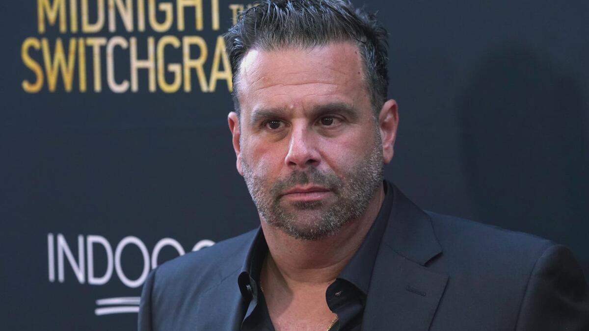 Man in a suit stands in front of a red carpet backdrop