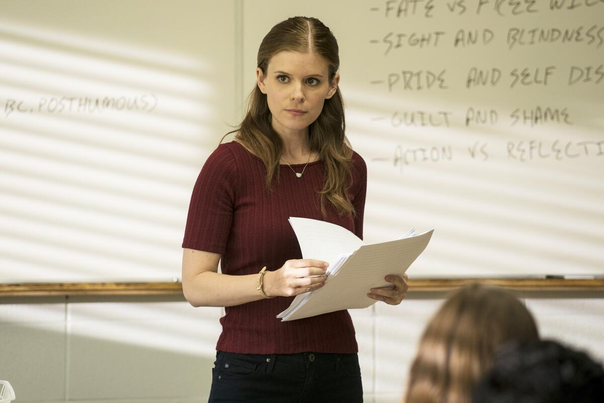 A teacher holding papers stands in front of a whiteboard