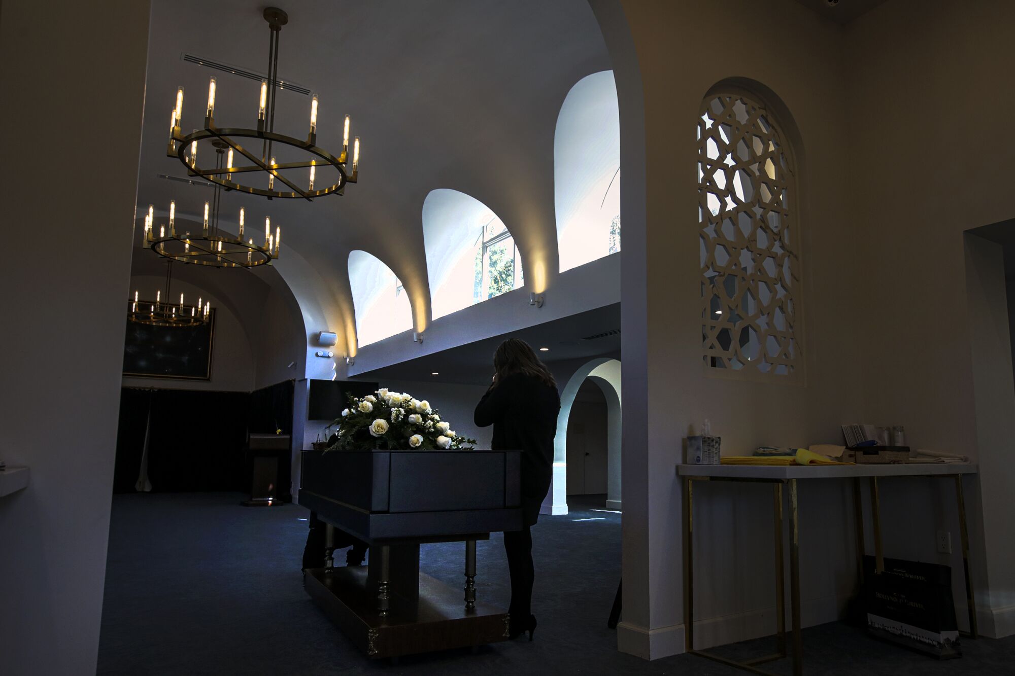 A lone woman is silhouetted next to a coffin topped with flowers in a dimly lit room with an arched ceiling