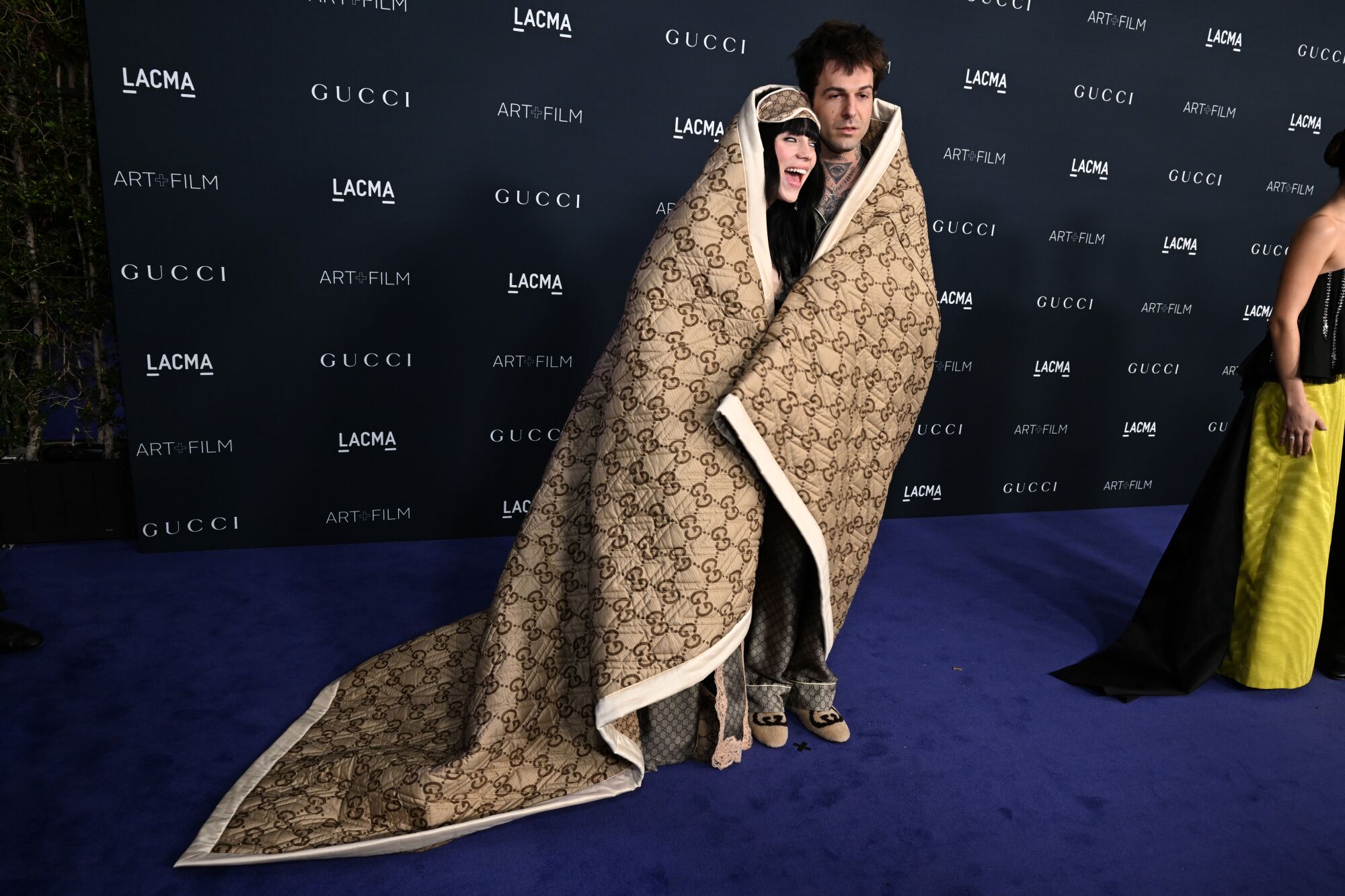 Billie Eilish and Jesse Rutherford wrapped in a blank and pose for a photo.