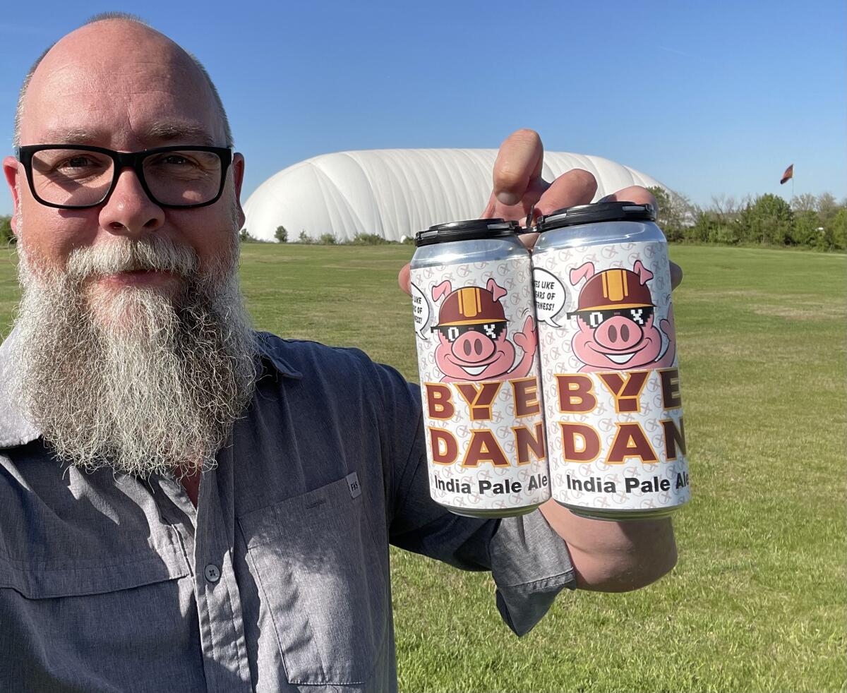 A bald man with a white bear and dark-rimmed glasses holds up two cars of beer with a cartoon pig on them on a grass field.