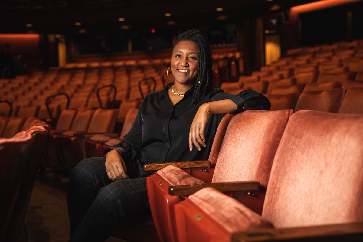 Miki Vale, seated and wearing black shirt and pants, gold jewelry, shoulder-length locs, poses inside The Old Globe theater