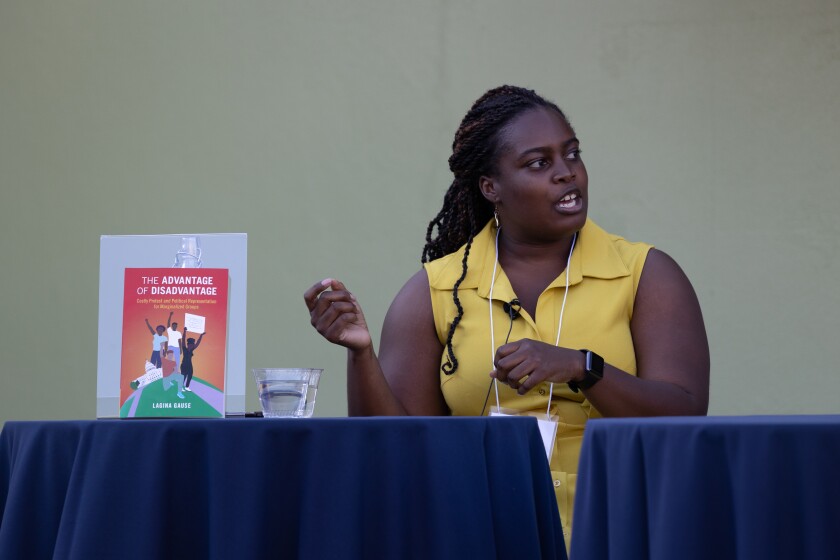 LaGina Gause shown from the waist up in a yellow dress, discussing her book, with a copy of the book propped up next to her