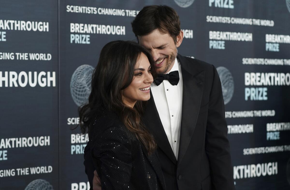 Mila Kunis smiles next to Ashton Kutcher, who is looking down at her and laughing, at a formal event