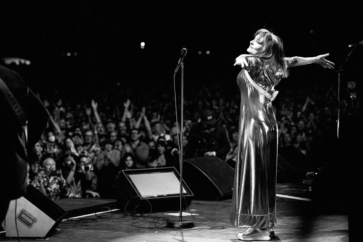 A woman stands onstage before a crowd, arms spread wide