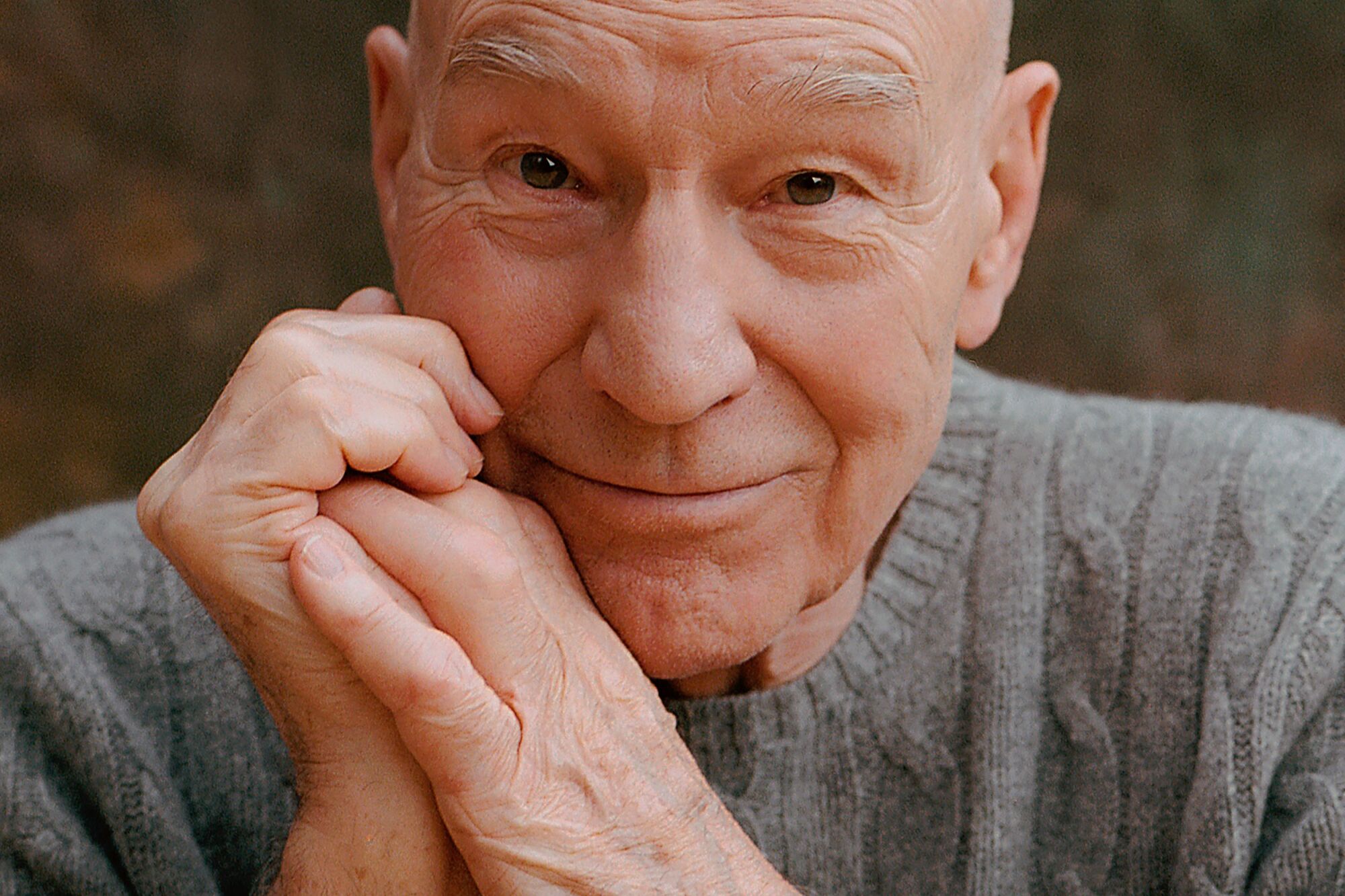 Patrick Stewart in a gray sweater with his arms clasped near his face.