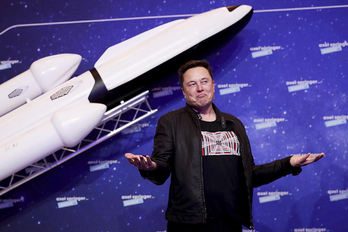 Elon Musk stands in front of a blue background and white rocket ship.