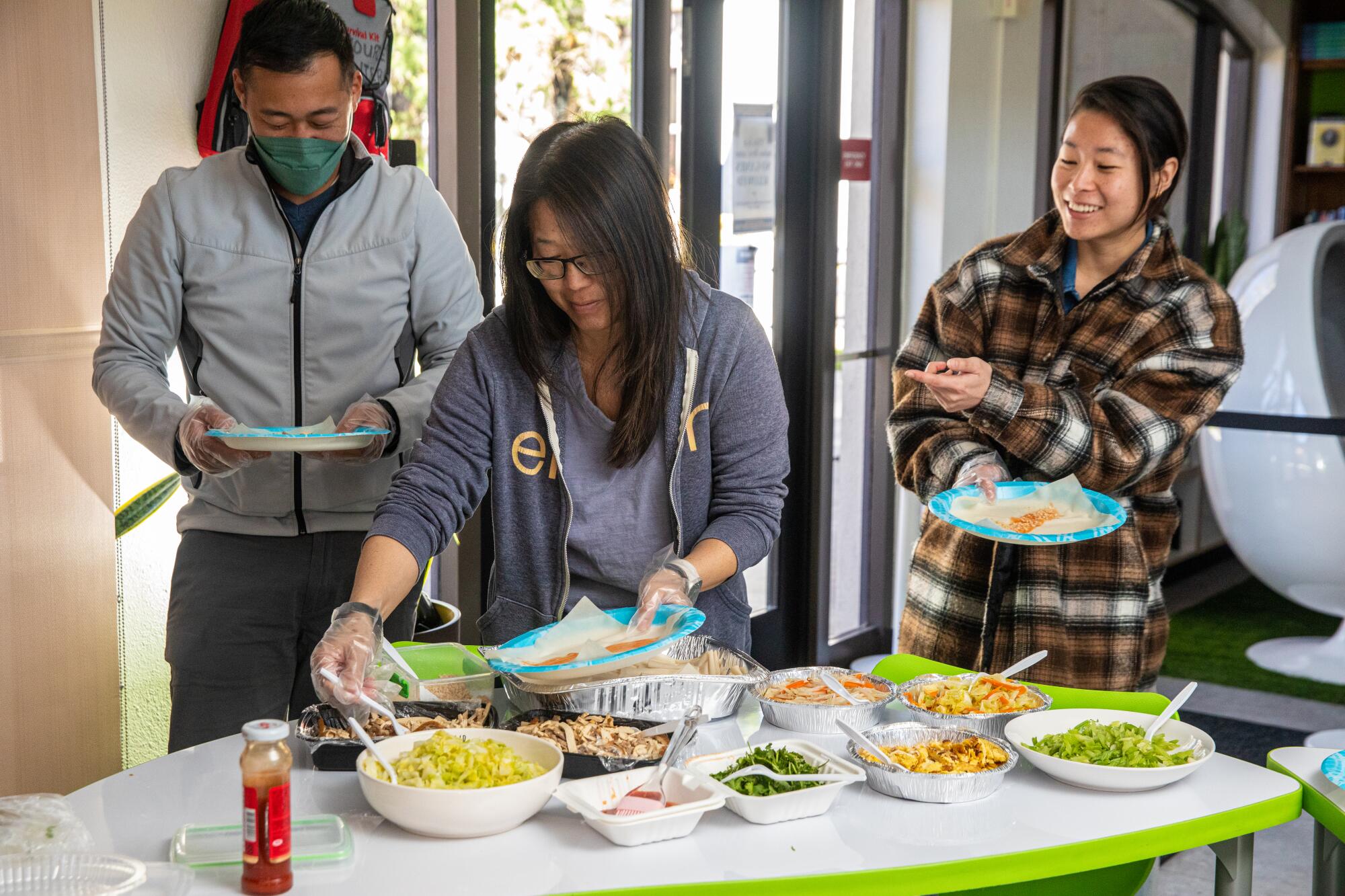 Three people stand around a table loaded with various plates of food items.