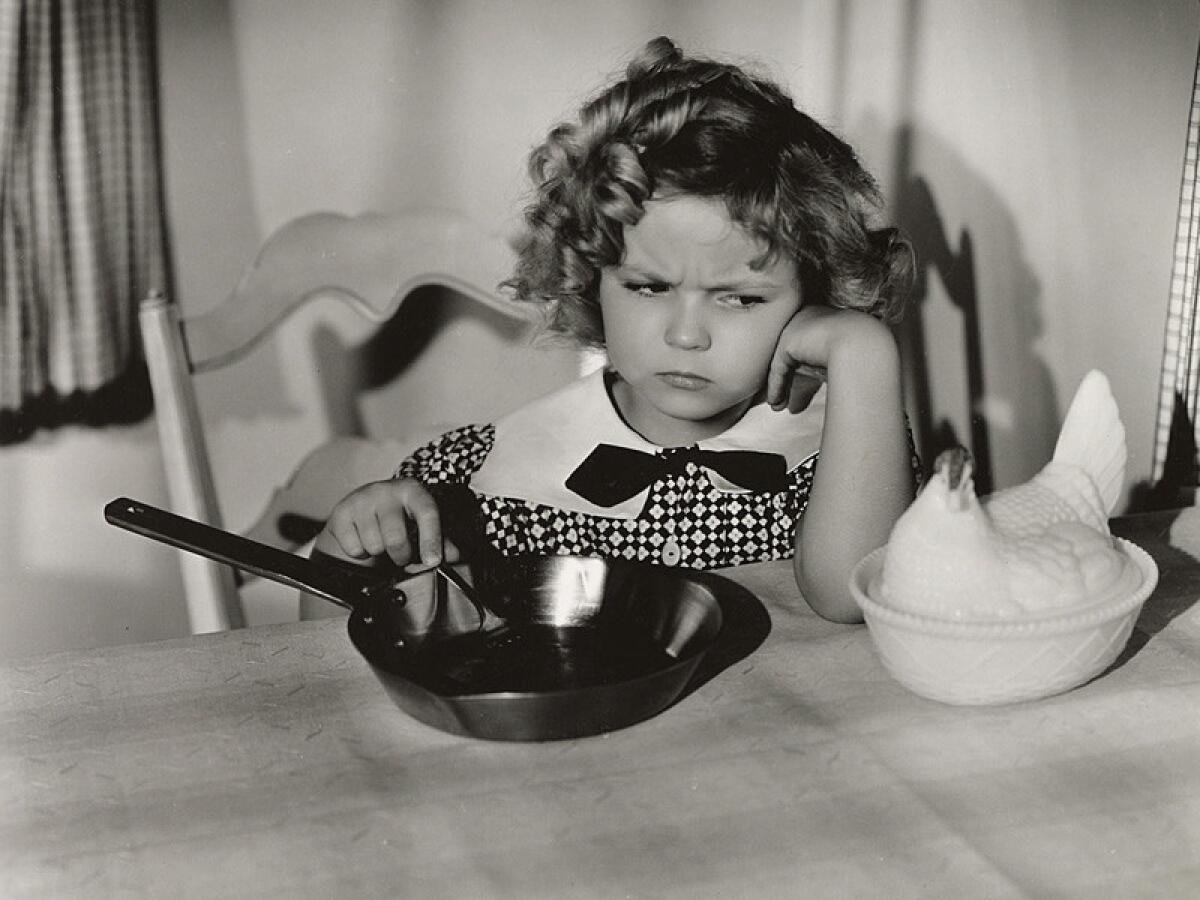 Fox star Shirley Temple trying to coax a china-dish hen to lay an egg so she can make an omelet, in the kitchen of Temple's bungalow cottage on the Fox studio lot, 1934.