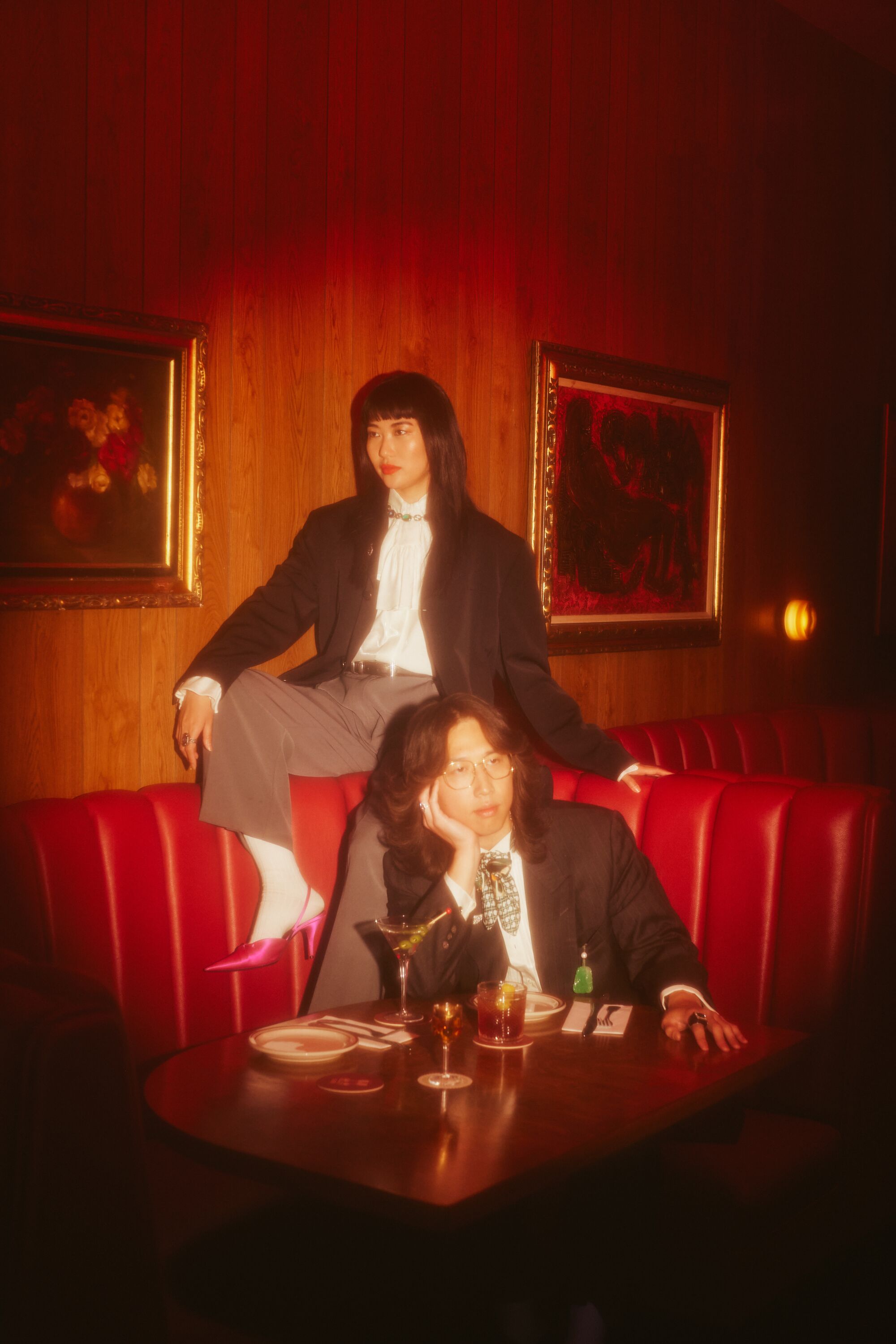 An Asian couple sitting in a restaurant with drinks, each looking to the side.  They wear vintage suits.