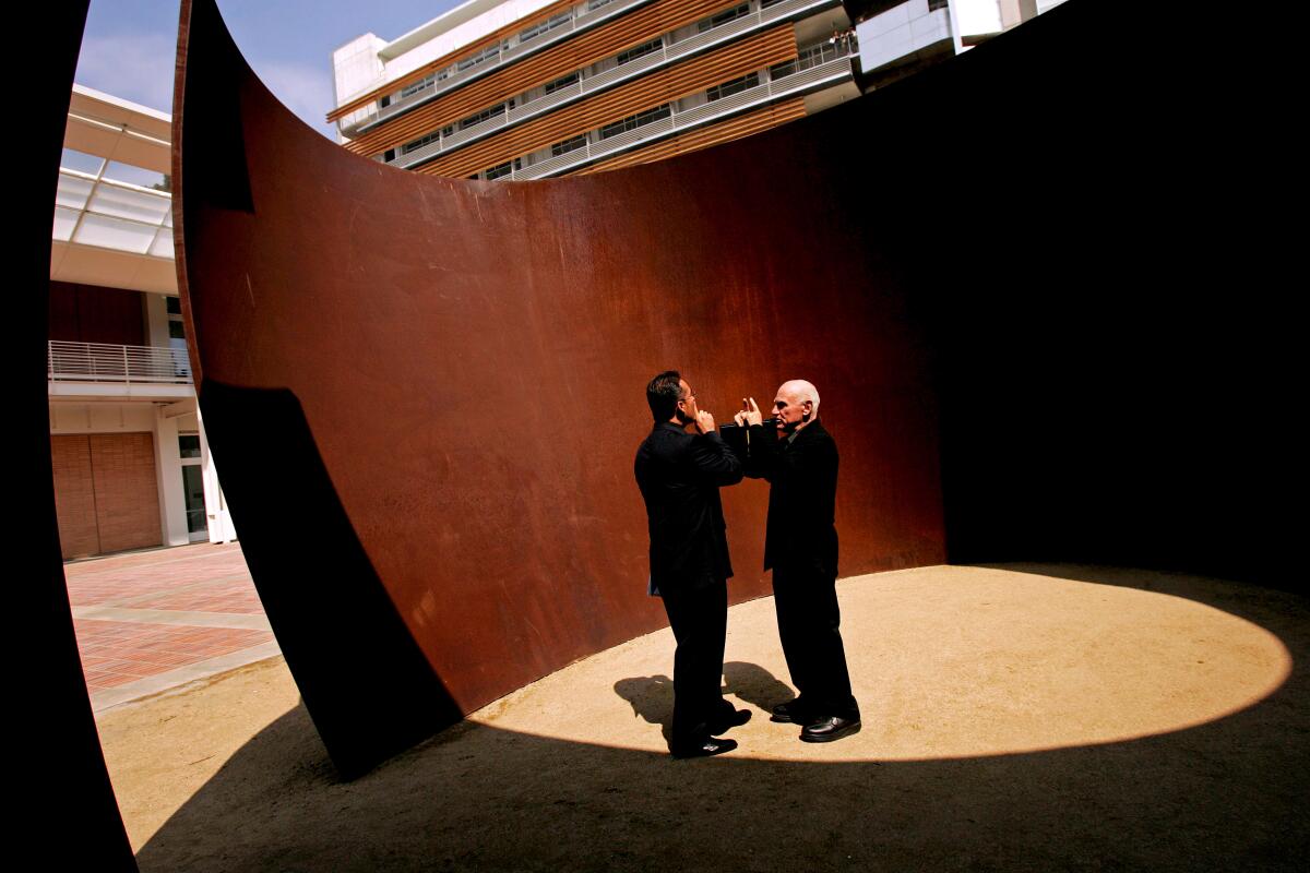 Richard Serra and Franciso Pinto, in black suits, stand inside a massive curved steel sculpture.