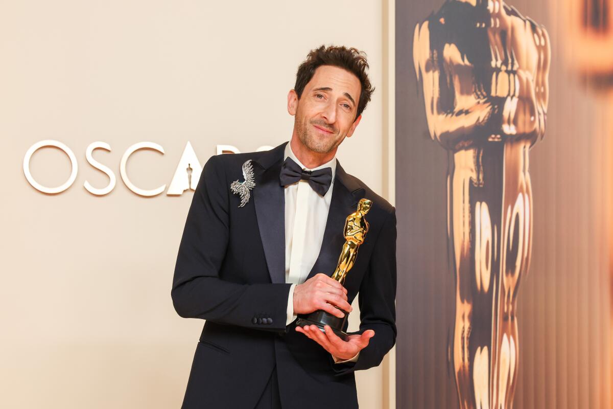 Adrien Brody in a tuxedo leans to his left and holds his Oscar statuette in his hands. He stands in front of a gold backdrop