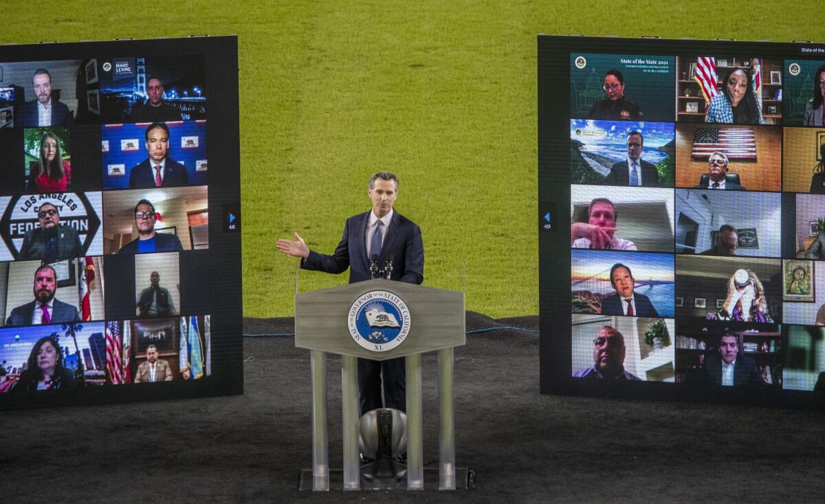 Gov. Gavin Newsom stands on a stage with large video boards on either side of him showing videoconference attendees