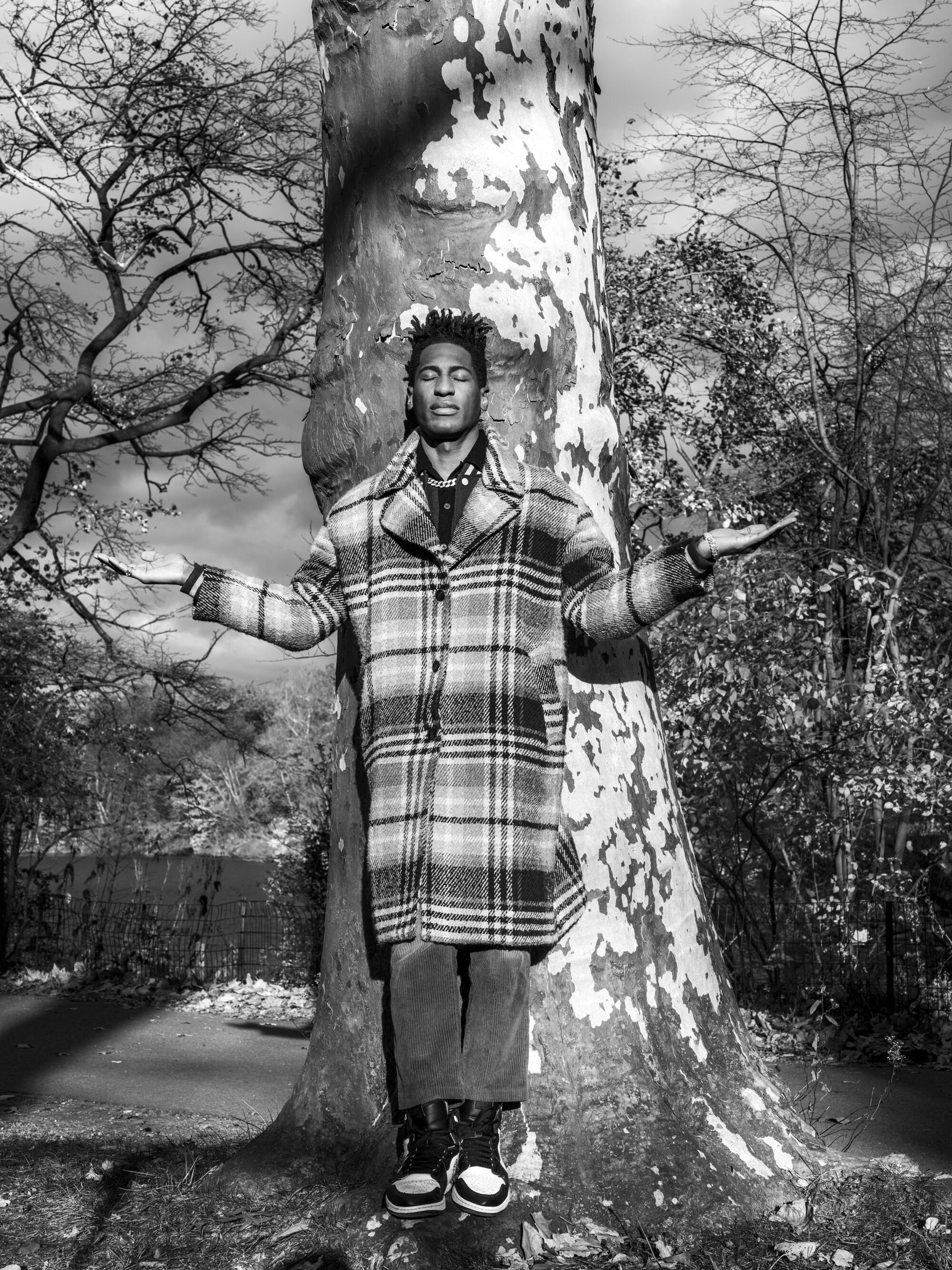 A man in a checkered coat leans against a tree and lifts his hands to his shoulders.