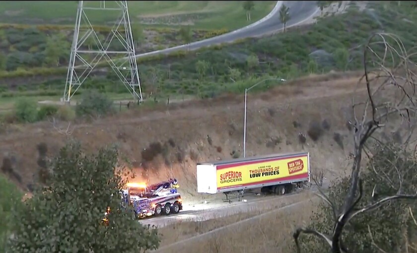 A tow truck is in front of a big rig's load on a road surrounded by grass and trees.