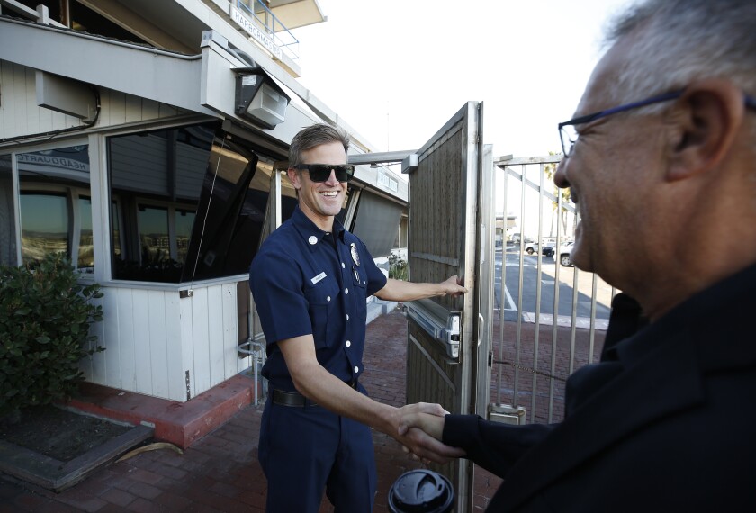 A man in a ship captain's uniform, left, holds open a gate door and shakes hands with a man in a black suit. 