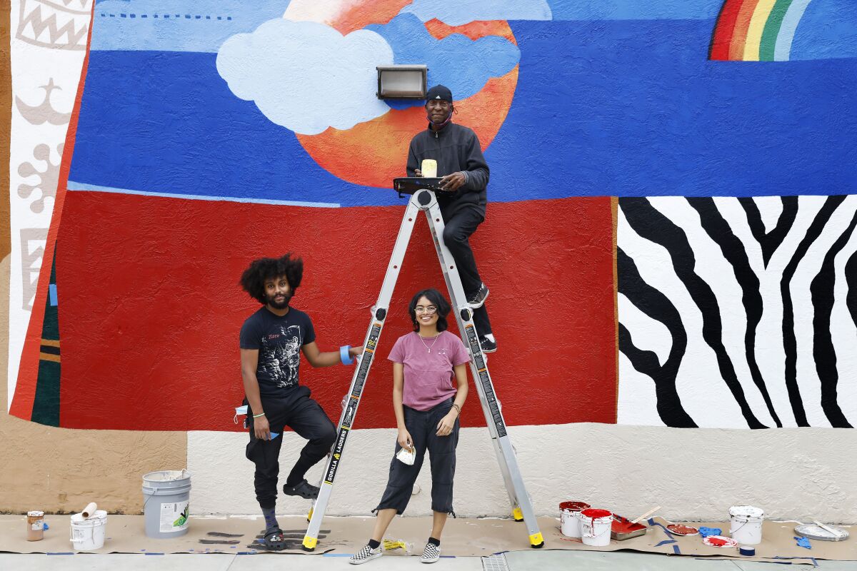 Two people below a ladder and one person on it in front of a mural