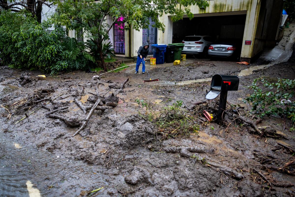 A man stands in a driveway with an open garage and trash cans. In the foreground are piles of debris and mud.