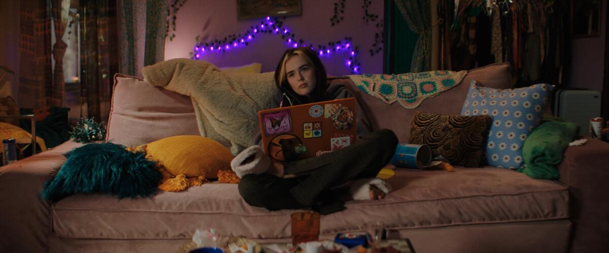 A young woman with a laptop sits on a couch in a darkened room.