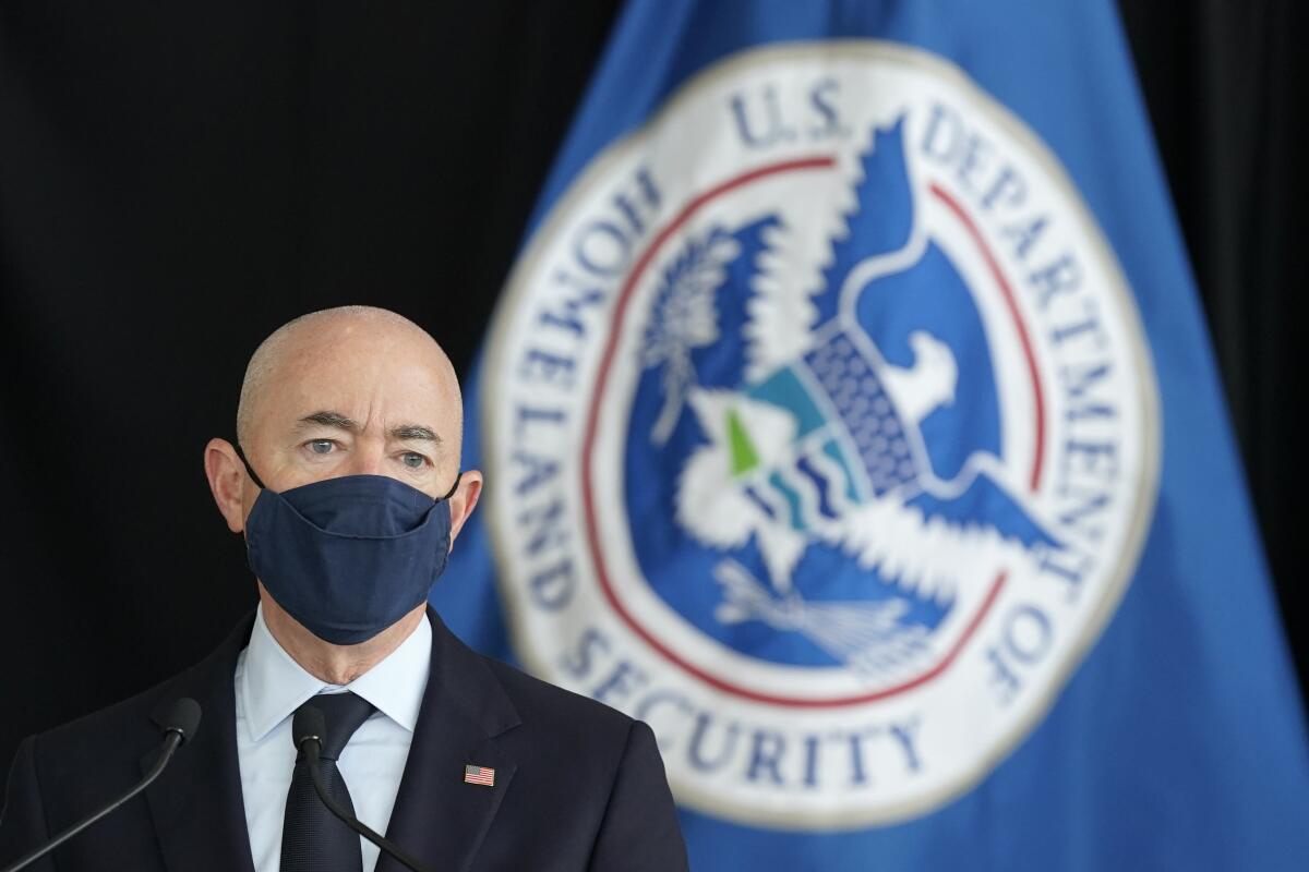 Alejandro N. Mayorkas wears a mask and stands in front of a Homeland Security Department flag