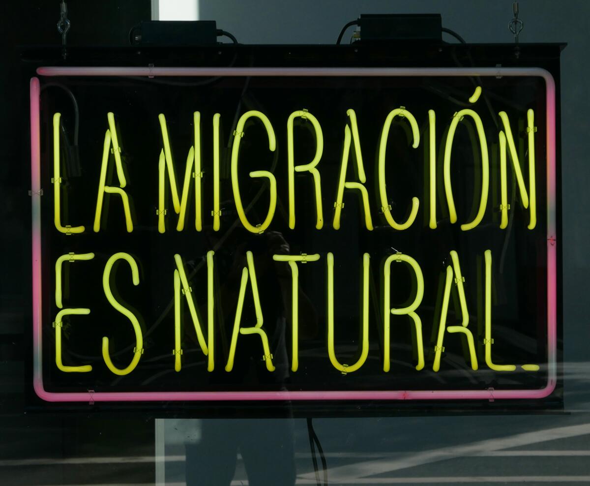 A neon sign that reads "La Migración es natural" is shown in a storefront window.