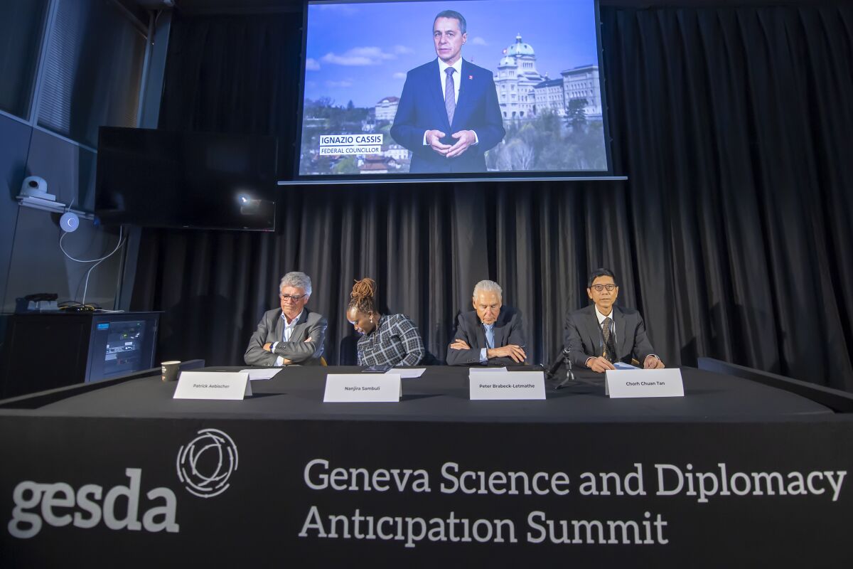 Patrick Aebischer, GESDA Vice Chairman, Nanjira Sambuli, Policy Analyst and Advocacy Strategist, Kenya, Peter Brabeck-Letmathe, GESDA Chairman, Chorh Chuan Tan, Chief Health Scientist, Singapore, left and right, and Swiss Federal Councilor Ignazio Cassis on the screen in recorded video, speaks about the first Geneva Science and Diplomacy Anticipation Summit 2021 (GESDA), during a press conference at the Campus Biotech in Geneva, Switzerland, Thursday, October 07, 2021. (Martial Trezzini/Keystone via AP)