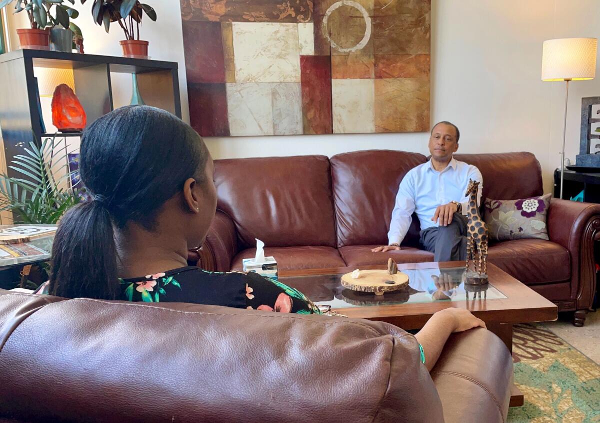 A man sits on a leather couch, speaking across a low table to a woman in a chair.