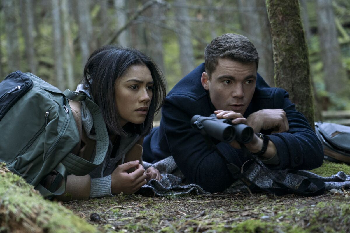 A woman, left, and a man holding binoculars are lying on the ground in a wooded area.