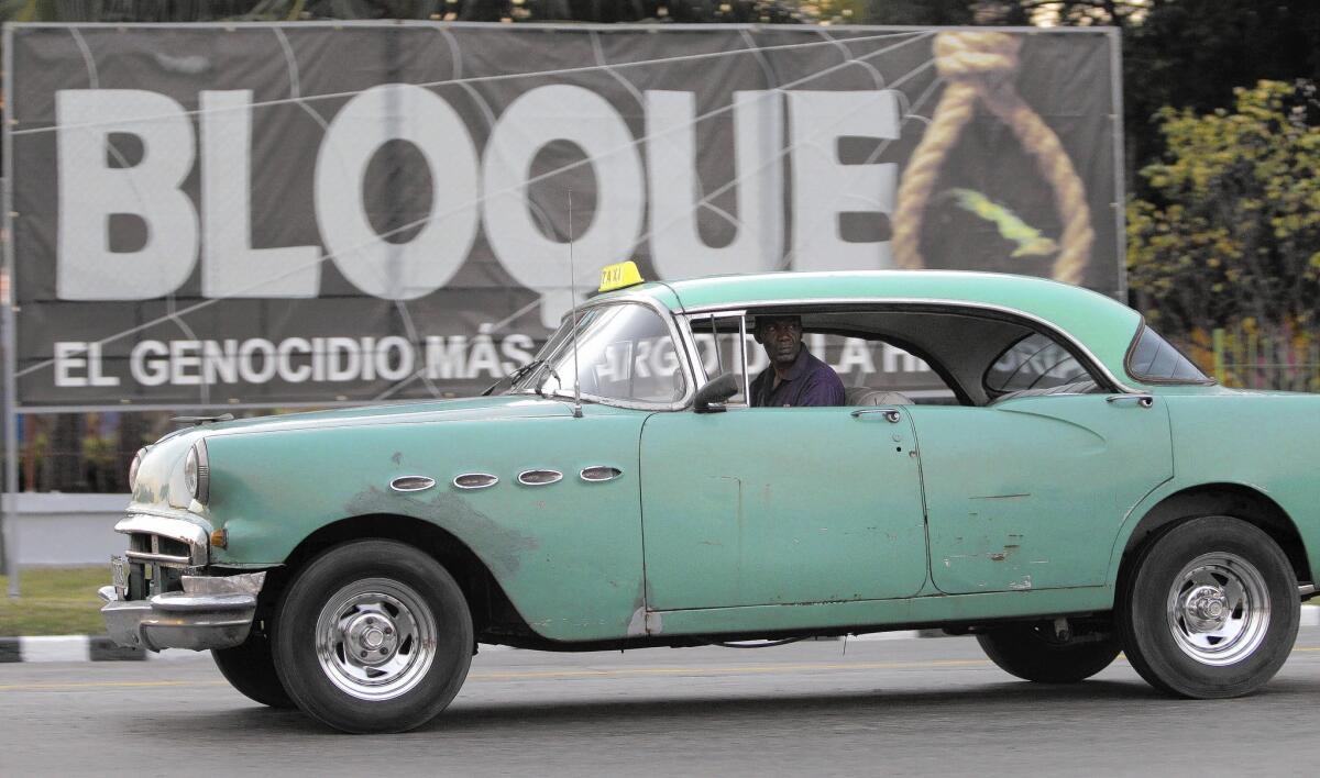 In Havana, a man drives a classic American car that serves as a taxi past a poster that reads in Spanish, "Embargo, the longest genocide of history."