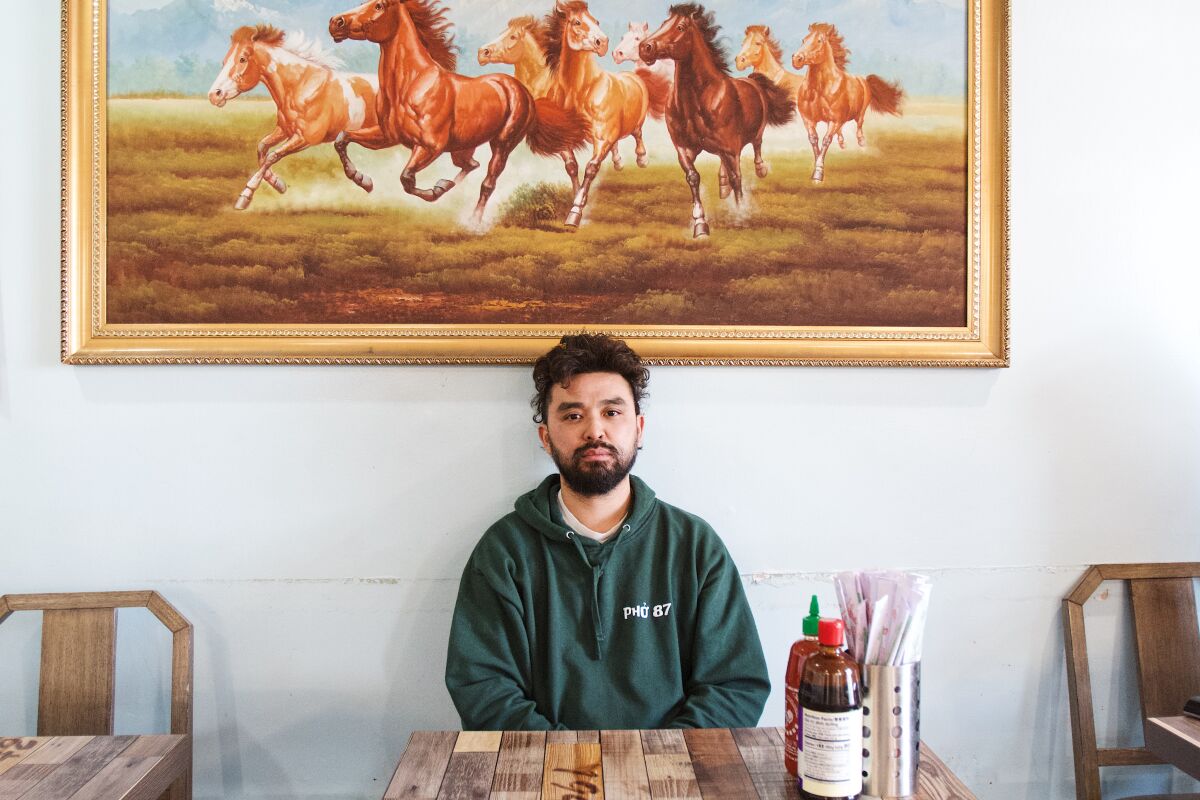 A horizontal portrait of Tre Dinh seated at one of his restaurant's tables. Behind him is a painting of wild horses.