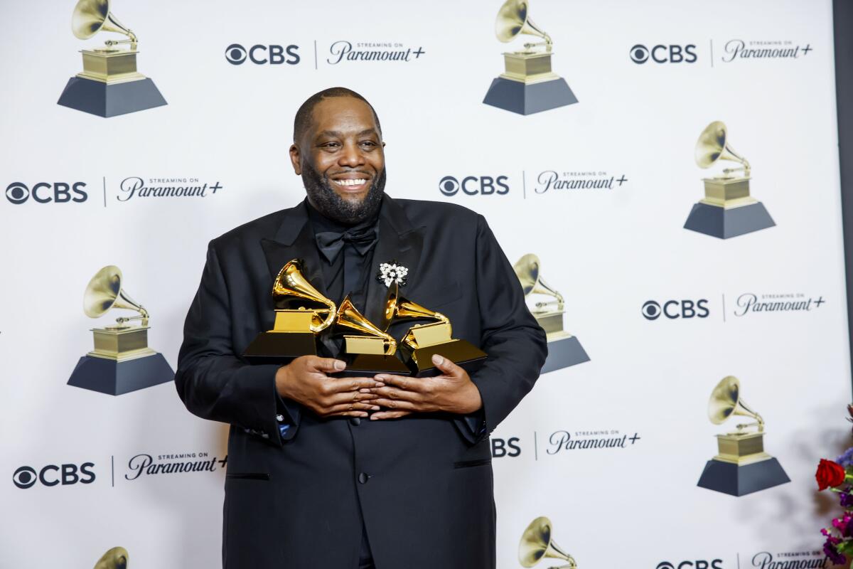 Killer Mike, clad in a black tuxedo and black shirt, smiles and cradles three Grammy Awards statues