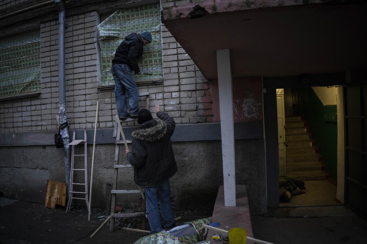 Two people cover the windows of a damaged building while another person lies in the building's doorway.