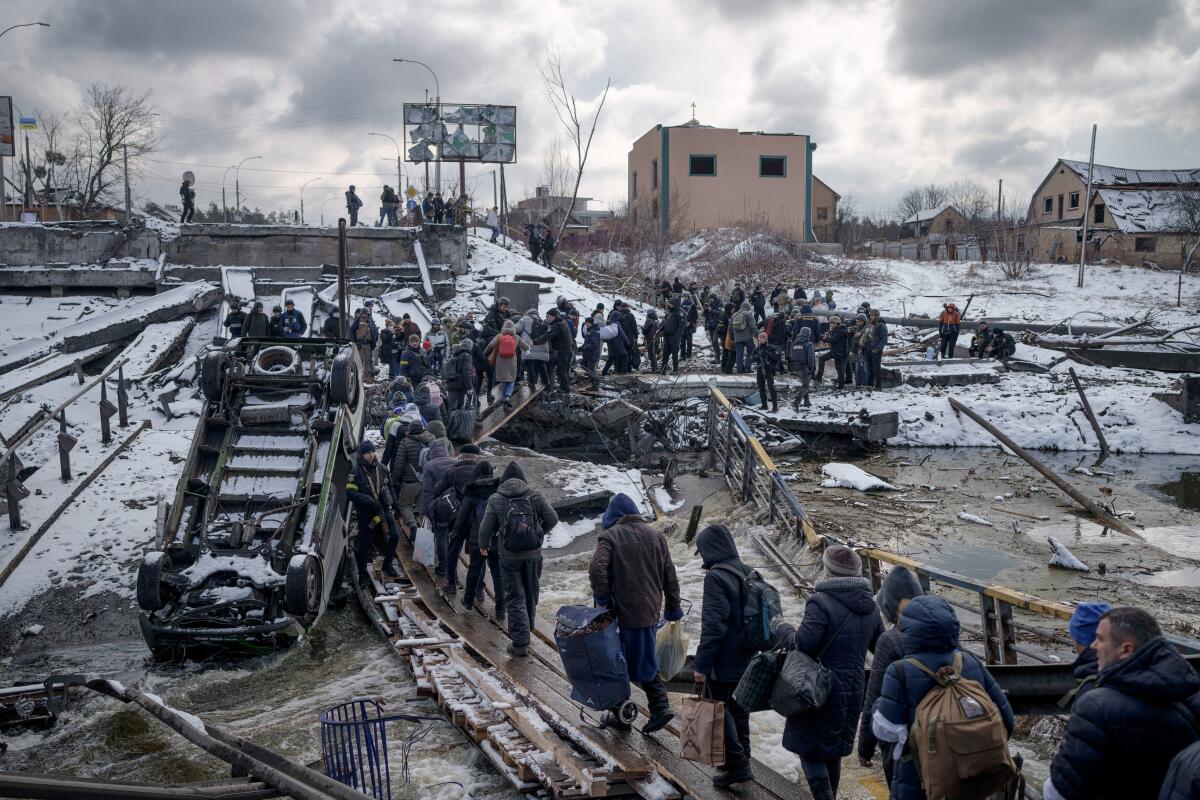 A long line of people carrying bags and walking on planks across a river 