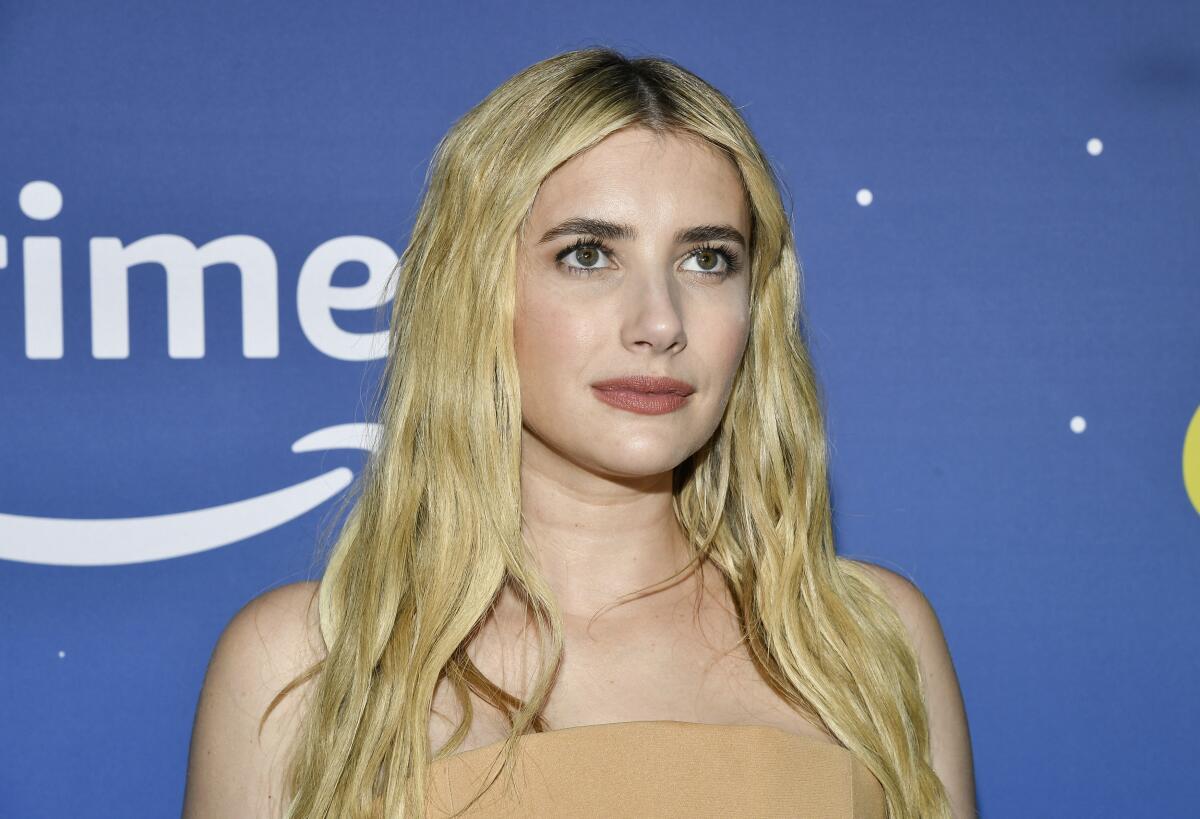 Emma Roberts gazes upward in a strapless dress while standing in front of a blue background.