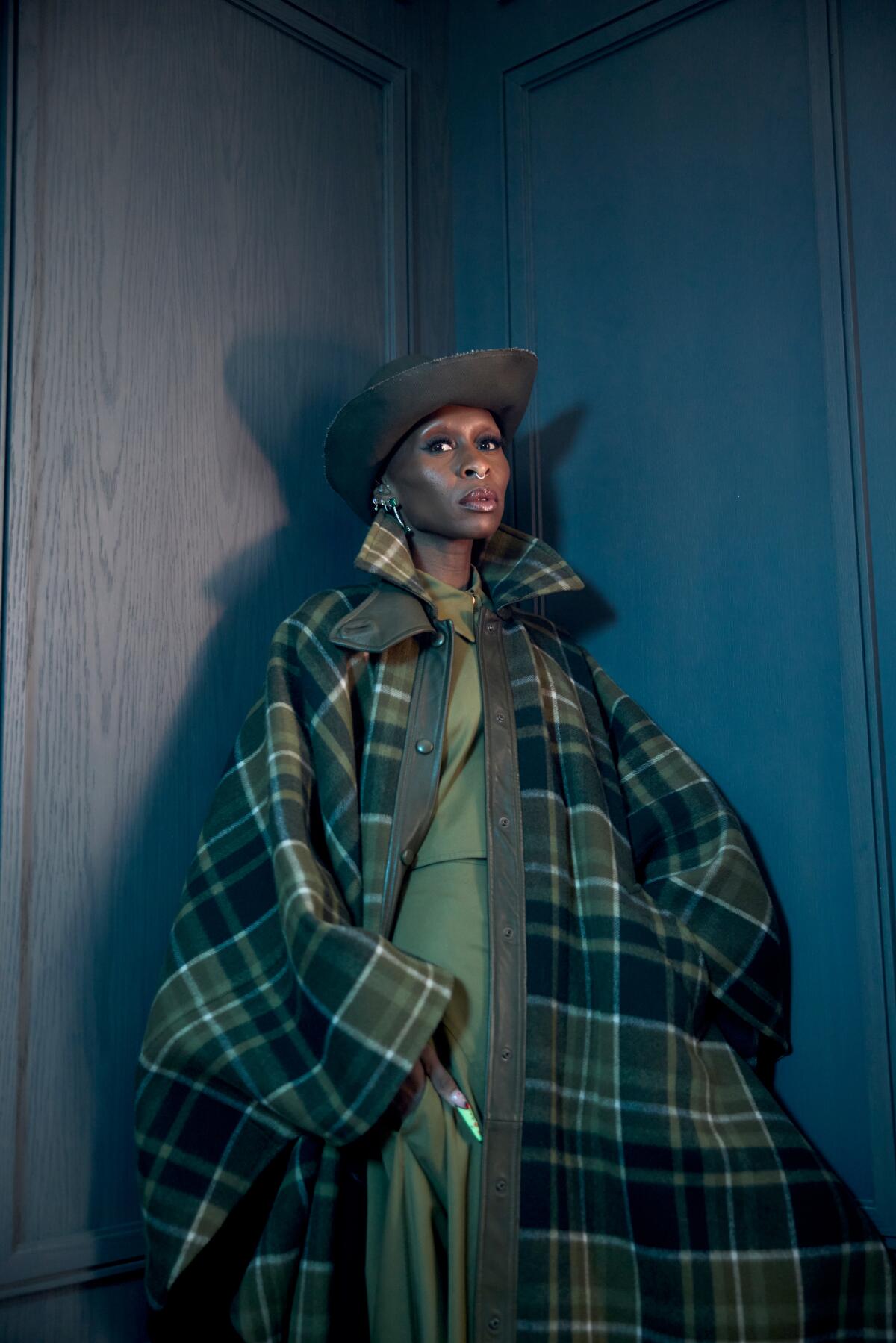 Cynthia Erivo stands in a corner, wearing a long plaid coat and jaunty hat